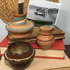 May include: Three small pottery pieces, a brown bowl, a brown and orange jar with a cork lid, and a taller brown and blue vase with a feather inside. The items are stacked on top of a stack of books.