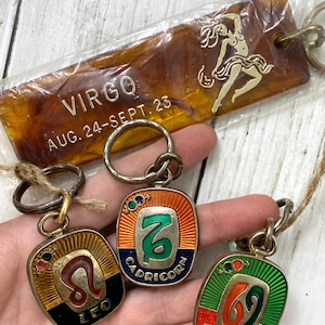 May include: A collection of vintage zodiac keychains. The keychains feature the Leo, Capricorn, and Cancer signs, with a Virgo keychain in the background. The keychains have a gold-tone metal finish with colorful enamel details. The Virgo keychain is a brown rectangle with gold text and a dancer illustration.