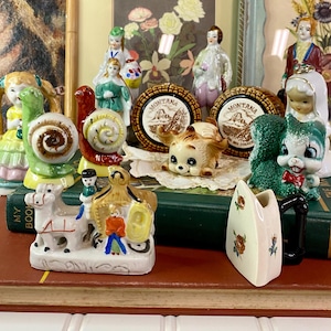 May include: A collection of vintage ceramic figurines, including a snail, a dog, a bride and groom, and a horse-drawn carriage. The figurines are in various colors and styles, with intricate details. Two small decorative plates with the word "Montana" are also visible.