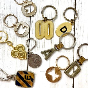 May include: A collection of vintage keychains in various shapes and designs. The keychains feature letters, numbers, and symbols in gold, silver, and brass tones. Some have intricate details, while others are simple and geometric.