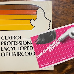 May include: A vintage Clairol Professional Encyclopedia of Haircolor book with a side profile illustration and a retro-style hair dryer. A pink booklet titled "The Chadwick System" is also present.