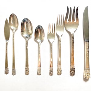 Puede incluir: Un surtido de cubiertos antiguos, que incluye cucharas, tenedores y un cuchillo, dispuestos sobre una superficie blanca. La cubertería presenta detalles florales ornamentados en los mangos y una superficie pulida y reflectante. El juego incluye cucharas y tenedores de varios tamaños.