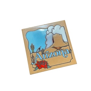 May include: A tan ceramic tile with a blue and white illustration of a desert landscape. The tile has the word "Arizona" written in blue cursive letters.