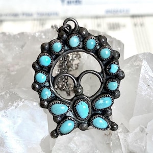 May include: A circular silver pendant featuring a series of small, round and oval turquoise cabochons set along the outer edge. The center of the pendant contains two symmetrical curved silver loops. A thin silver chain is attached to the top loop.