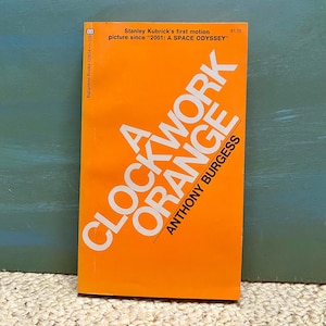 May include: A Clockwork Orange book cover with an orange background and white text. The title "A Clockwork Orange" is in large, bold letters. The author's name, "Anthony Burgess", is in smaller letters below the title.