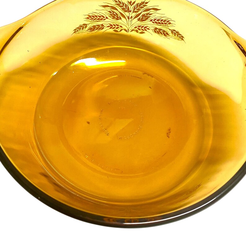 K&ouml;nnte beinhalten: Bernsteinfarbene Pyrex-Auflaufform mit Weizenmuster. Die Sch&uuml;ssel hat eine runde Form mit zwei Griffen und eine transparente Glaskonstruktion. Das Weizenmuster ist braun und befindet sich auf der Oberseite der Sch&uuml;ssel.