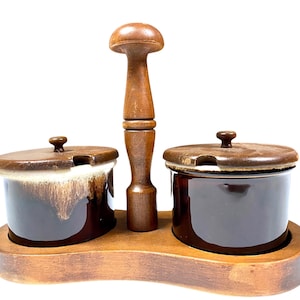 May include: A vintage brown ceramic condiment set with two small lidded jars and a wooden handle, all set on a wooden tray. The jars have a dark brown glaze with a lighter, dripped effect. The wooden handle is tall and mushroom-shaped.