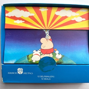 May include: A blue box containing 12 self-mailers and 12 seals from American Greetings. The box features a cartoon illustration of a character, a dog, and a duck on a globe under a yellow and red sunburst sky with white clouds and a hot air balloon.