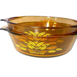 K&ouml;nnte beinhalten: Zwei bernsteinfarbene, oval geformte Glasschalen. Die Schalen sind gestapelt und haben an jeder Seite Griffe. Ein goldenes Weizenmuster verziert die Vorderseite der Schalen. Die Schalen dienen wahrscheinlich zum Servieren von Speisen.