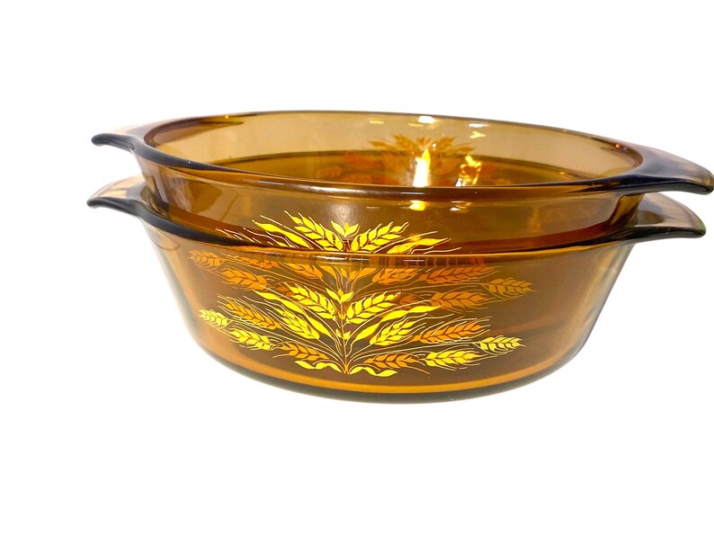 K&ouml;nnte beinhalten: Zwei bernsteinfarbene, oval geformte Glasschalen. Die Schalen sind gestapelt und haben an jeder Seite Griffe. Ein goldenes Weizenmuster verziert die Vorderseite der Schalen. Die Schalen dienen wahrscheinlich zum Servieren von Speisen.