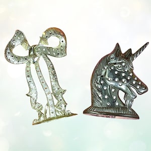 May include: Two silver decorative items: a bow and a unicorn head. The bow has intricate detailing and is adorned with small, sparkling accents. The unicorn head has a detailed mane and a spiralled horn, also embellished with sparkling accents.