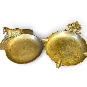 May include: Two brass trays. The left tray is circular with a smooth rim and a sculpted horse figure resting on one side. The right tray is round with a scalloped edge, three integrated cigarette rests, and a decorative floral motif on the rim.