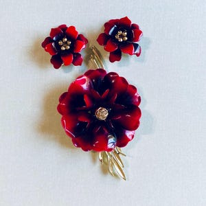May include: A set of three red and gold flower-shaped brooches. The largest brooch has a single flower with a gold center and leaves. The two smaller brooches are earrings with a single flower each.