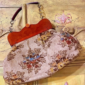 May include: Vintage handbag with a floral pattern in shades of brown, yellow, and blue on a beige background. The bag features a reddish-orange frame, a chain strap, and a dark brown handle. A round, orange clasp secures the bag.