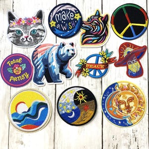 Puede incluir: Una colección de parches bordados con varios diseños. Incluye un gato con una corona de flores, un lobo, un oso, un símbolo de la paz, una seta, un símbolo del yin-yang, un sol y una luna, y parches con las frases "make a wish" y "Total Pansy".