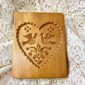 May include: A rectangular light brown wooden cookie mold featuring a carved heart shape with a scalloped border. Inside the heart, two birds face each other above a floral stem, with small four-petaled flower motifs positioned above the birds.