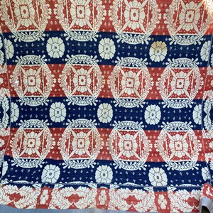 May include: A vintage quilt with a repeating pattern of red and white floral medallions and blue stripes. The quilt has a light blue border and is likely made of cotton or a similar fabric. The design is symmetrical and features detailed floral elements.