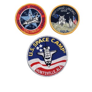May include: Three embroidered patches featuring space-themed designs. The first patch is red, white and blue with a space shuttle and the text "GRAND OVERMYER ALLEN COLUMBIA LENOR". The second patch is blue with a white star and the text "FIRST LUNAR LANDING 1969 USA". The third patch is blue with a white star and the text "U.S. SPACE CAMP HUNTSVILLE, ALA."