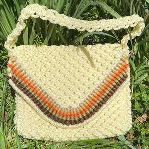 May include: A crocheted bag with a cream-colored body and a brown, orange, and green striped trim. The bag has a long strap and is made of a soft, textured yarn.