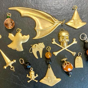Puede incluir: Una colección de abalorios y cuentas doradas con temática de Halloween. Los abalorios incluyen un ala de murciélago, sombreros de bruja, una calavera y huesos cruzados, un fantasma, gatos, una espada y una lápida. Algunos abalorios se combinan con cuentas decorativas.