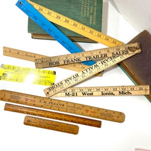 May include: A collection of vintage wooden and plastic rulers in various colors and sizes, some with printed text. The rulers are arranged on a white surface, with a green book in the background. Some rulers have measurements up to 35 inches.