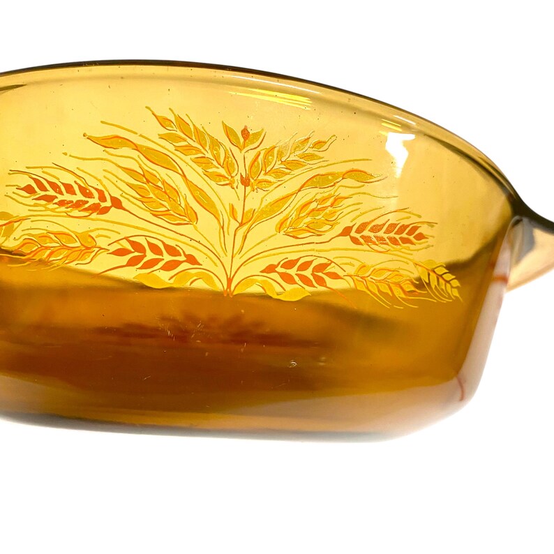 K&ouml;nnte beinhalten: Bernsteinfarbene Backform aus Glas mit einem Weizen&auml;hren-Design. Die Form hat eine rechteckige Form mit abgerundeten Ecken und einem Griff auf einer Seite. Das Weizendesign ist in Gelb- und Oranget&ouml;nen gehalten.