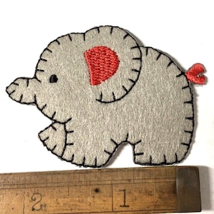 May include: A grey felt elephant applique with black stitched details and a red ear and tail. The elephant is approximately 5 cm long. The applique is on a white surface.