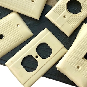 Puede incluir: Una colección de tapas de tomacorrientes y interruptores de luz vintage de color marfil. Las tapas tienen una textura acanalada y están hechas de baquelita.