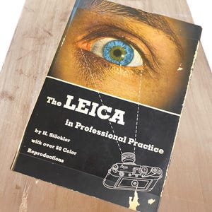 Puede incluir: La portada de un libro titulado "The Leica in Professional Practice" de H. St"ockler. La portada presenta un primer plano de un ojo azul y un dibujo lineal de una cámara Leica.