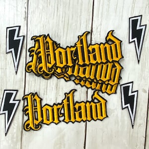 May include: Embroidered patches with the word "Portland" in a gothic font, in yellow thread on a black background. The patches are surrounded by black and white lightning bolt patches. The patches are on a light wood grain surface.