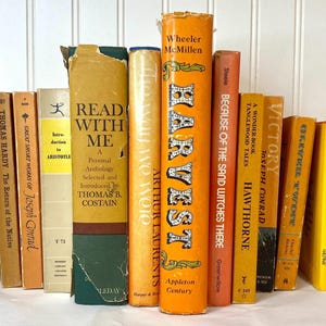 May include: A collection of vintage books with various titles and colors. The spines are primarily orange, yellow, and tan, with titles like "Harvest," "Victory," and "Read With Me." The books are arranged upright, suggesting a library or decorative display.