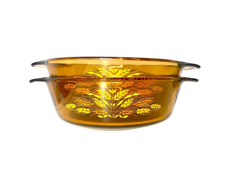 K&ouml;nnte beinhalten: Zwei bernsteinfarbene Auflaufformen aus Glas mit einem Weizen&auml;hren-Design in Gelb- und Oranget&ouml;nen. Jede Form hat schwarze Griffe. Die Formen sind gestapelt. Ma&szlig;e in Zentimetern.