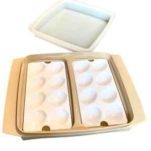 May include: A beige plastic egg container with a lid. The container has two sections, each holding six white egg-shaped molds. The lid is a square shape and is a lighter shade of beige.