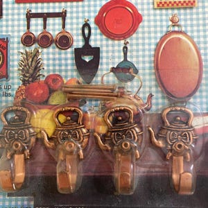May include: A package of four bronze-colored teapot-shaped wall hooks. The hooks are in a clear plastic package, with a variety of kitchen-themed decorative items in the background, including measuring spoons, a spatula, and a tray.