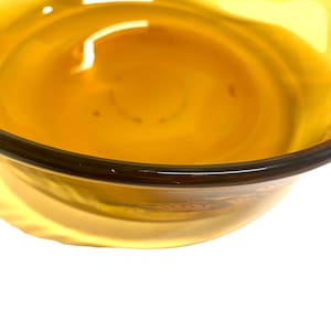 K&ouml;nnte beinhalten: Nahaufnahme einer klaren, bernsteinfarbenen Glasschale. Die Schale hat einen dunkelbraunen Rand und eine glatte, reflektierende Oberfl&auml;che. Das Innere der Schale ist mit einem helleren Bernsteinton gef&uuml;llt, wodurch ein Gradienteneffekt entsteht. Die Schale steht vor einem wei&szlig;en Hintergrund.