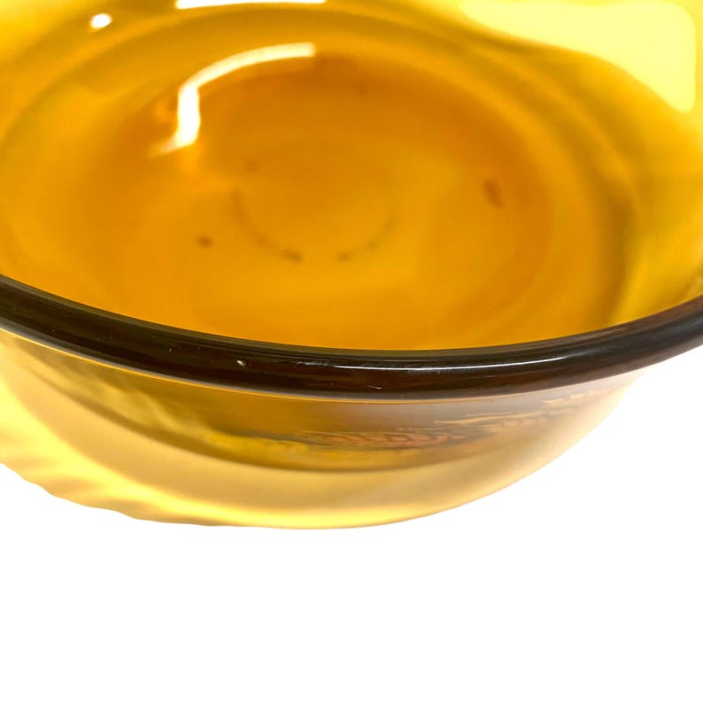 K&ouml;nnte beinhalten: Nahaufnahme einer klaren, bernsteinfarbenen Glasschale. Die Schale hat einen dunkelbraunen Rand und eine glatte, reflektierende Oberfl&auml;che. Das Innere der Schale ist mit einem helleren Bernsteinton gef&uuml;llt, wodurch ein Gradienteneffekt entsteht. Die Schale steht vor einem wei&szlig;en Hintergrund.