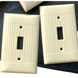 Puede incluir: Tres placas de interruptor de luz de plástico de color crema con ranuras verticales. Cada placa tiene un solo interruptor de palanca.