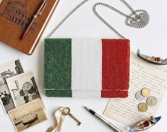 May include: A beaded clutch with the Italian flag design in green, white, and red. The rectangular bag has a silver chain strap. The image also includes a brown notebook, postcards, a key, a pen, and coins.