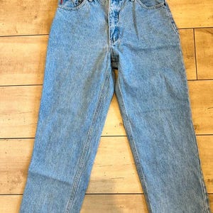 May include: High-waisted light blue denim jeans featuring a classic five-pocket design, a button fly closure, and a straight-leg cut. The denim has a medium wash with visible stitching details along the seams and a small red brand tag on the pocket.