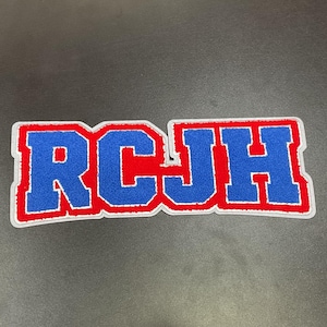 May include: A rectangular patch with the letters "RCJH" in blue, outlined in red and white. The patch has a felt-like texture and is likely for a jacket or bag. The background is a solid dark gray.