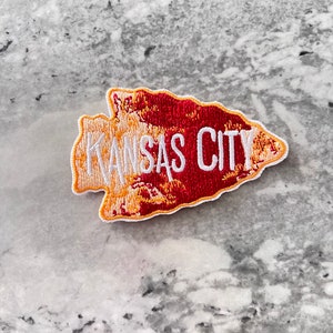 May include: A red and yellow embroidered patch shaped like an arrowhead with the text "KANSAS CITY" in white.