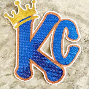 May include: A blue and gold sequined patch with the letters "KC" and a gold crown on top. The patch is outlined in orange and white.