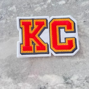 May include: White felt patch with the letters "KC" in red and yellow outlined embroidery.