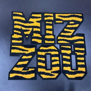 May include: Embroidered patches spelling out "MIZ ZOU" in a bold, block letter style. The letters feature a yellow and black tiger stripe pattern with a black border, perfect for apparel or accessories.