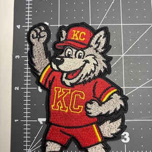 May include: Embroidered patch featuring a cartoon wolf in a red uniform with yellow accents. The wolf is wearing a red cap with "KC" in yellow. The patch is on a black surface with inch markings.