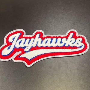 May include: A red, white, and blue patch with the word "Jayhawks" in a script font. The text is outlined in white and has a red border. The patch is likely for apparel or accessories.