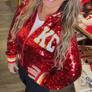 May include: A red sequined bomber jacket with the letters "KC" in white and yellow. The jacket has a zipper closure and striped cuffs in red, yellow, and white. The jacket is worn with black leggings.