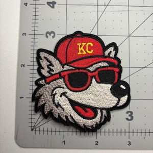 May include: Embroidered patch featuring a cartoon wolf wearing a red baseball cap with "KC" in yellow, and red sunglasses. The wolf has gray and white fur, a black outline, and a smiling expression. The patch is approximately 3 inches wide.