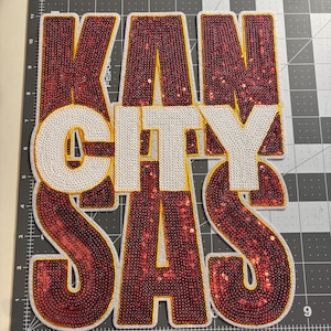 May include: A large, embroidered patch with the words "KANSAS CITY" in a bold, stacked design. The letters "KAN SAS" are in red sequins with a gold outline, while "CITY" is in white. The patch is on a gray cutting mat.