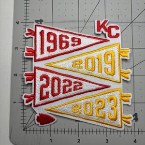 May include: Embroidered patch featuring pennant flags with the years 1969, 2019, 2022, and 2023 in red and gold. The patch includes the letters "KC" in red. The pennants are white with red and gold trim.